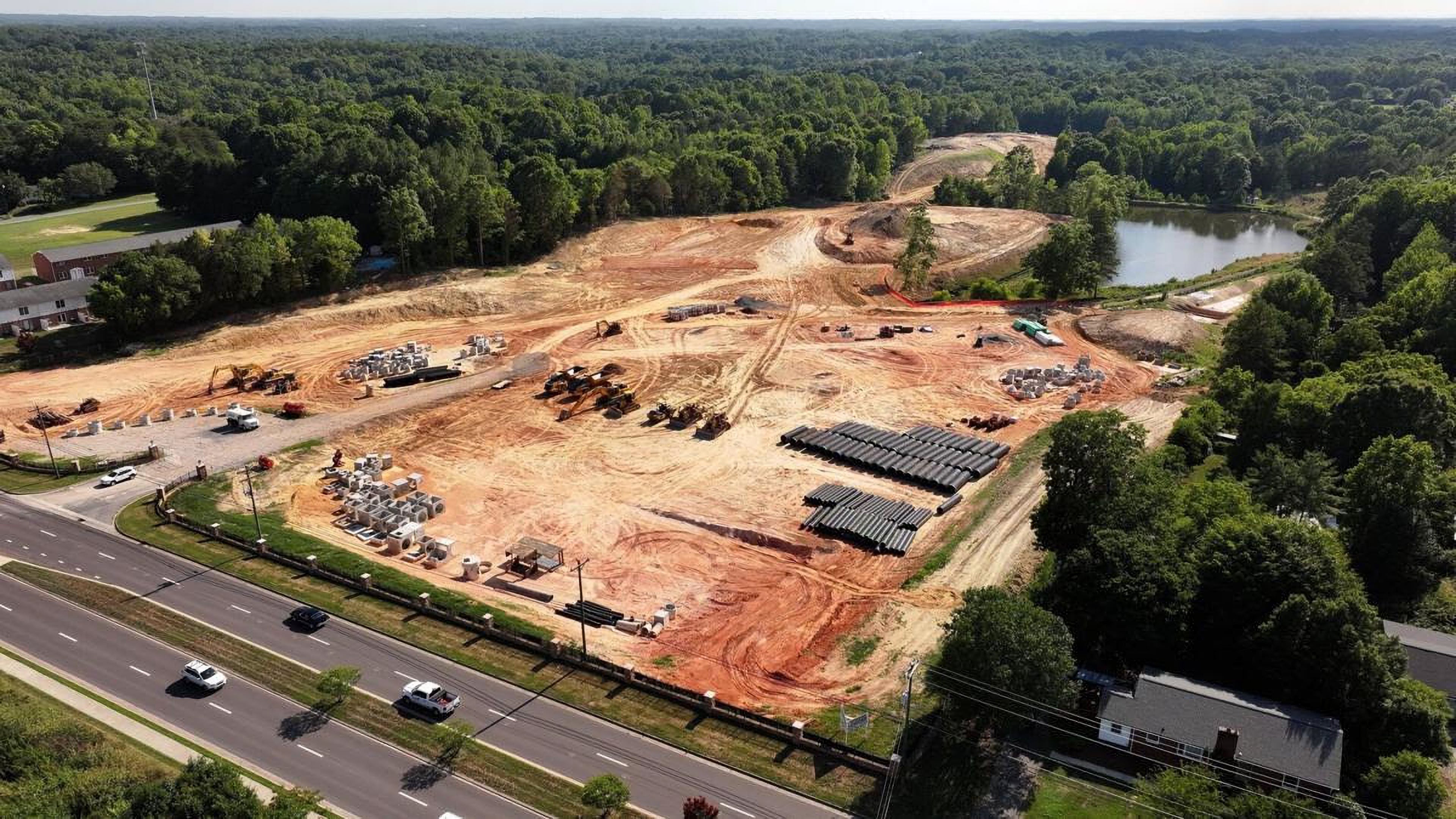 Aerial view of The Lake at Belmont's site under construction: cleared and graded red clay, earthmovers, stacked drainage pipe and building materials, with a lake behind the site and a four-lane road in front