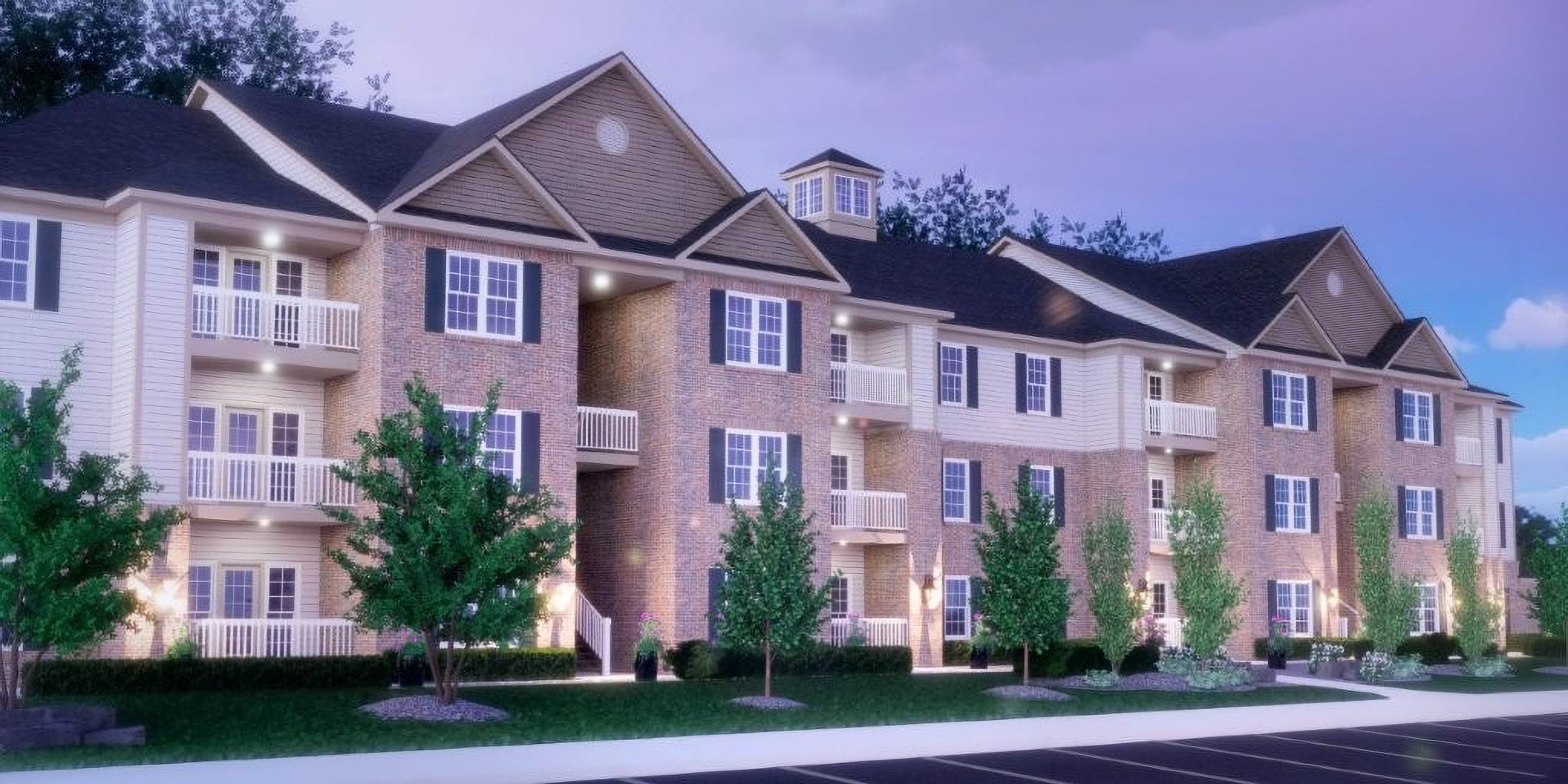 Rendering of The Lake at Belmont at dusk: three-story apartment buildings in brick and pale siding, with white balconies and lit entries, behind a lawn, young trees and a parking lot